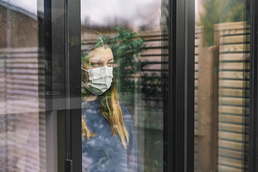 A woman wearing a mask, looking out the window during the COVID-19 pandemic