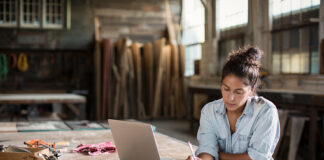 Mixed-race young entrepreneur with dark hair wears light blue chambray shirt with sleeves rolled up to her elbows. She holds a pencil while sketching or doing math in her paper notebook. Her laptop is adjacent, as is a smart phone. Also visible in the foreground are carpentry tools and sawdust. Natural light in the lofty, spacious maker space illuminates industrial tools and raw lumber in a soft-focus background. She has the confidence and energy to solve any design challenge and to deliver the prototype on time and under budget.