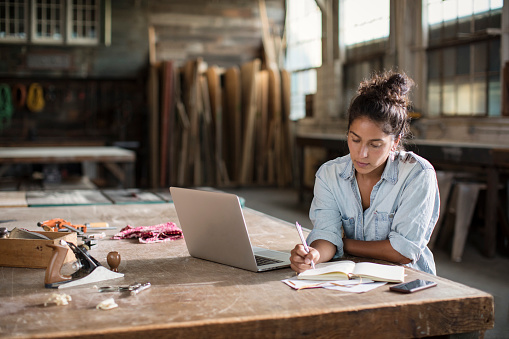 Mixed-race young entrepreneur with dark hair wears light blue chambray shirt with sleeves rolled up to her elbows. She holds a pencil while sketching or doing math in her paper notebook. Her laptop is adjacent, as is a smart phone. Also visible in the foreground are carpentry tools and sawdust. Natural light in the lofty, spacious maker space illuminates industrial tools and raw lumber in a soft-focus background. She has the confidence and energy to solve any design challenge and to deliver the prototype on time and under budget.